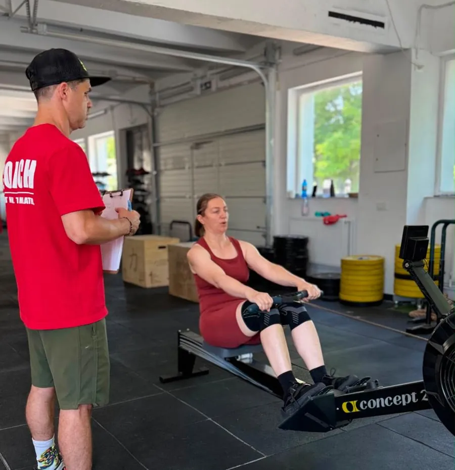 Antrenor CrossFit C15 în tricou roșu, cu clipboard, urmărind o membră care se antrenează la aparatul de canotaj Concept2