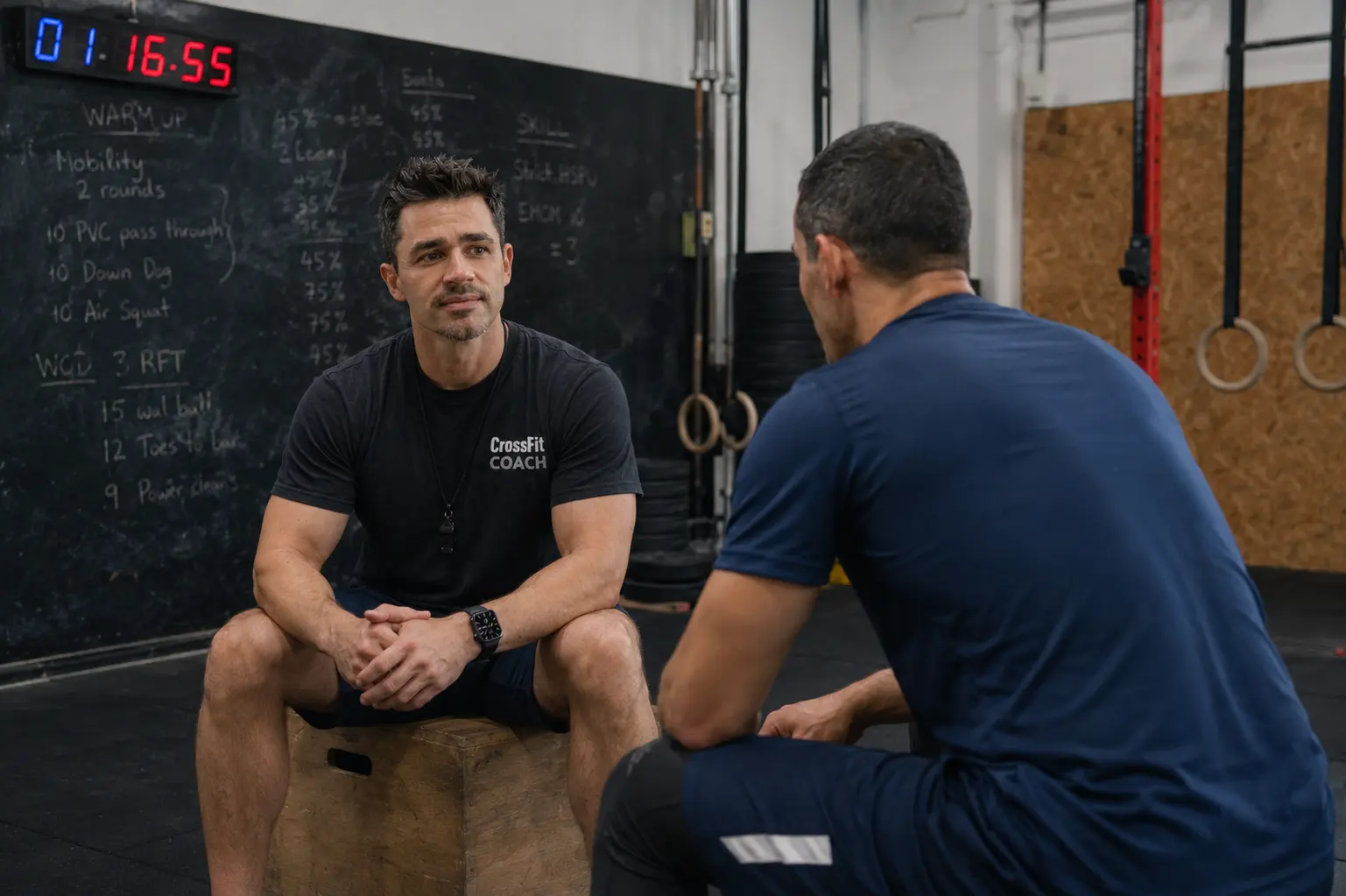Un antrenor CrossFit C15 discutând față în față cu un membru, așezați pe cutii, cu tabla de antrenament în fundal